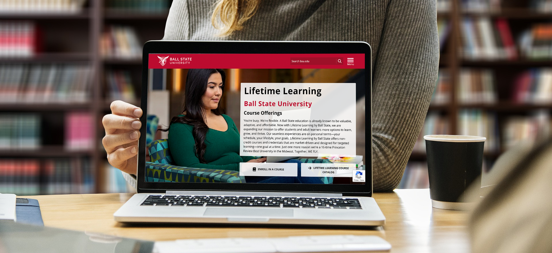 A woman in a gray sweater sits in a library and holds up a laptop with the Lifetime Learning homepage loaded on the screen.