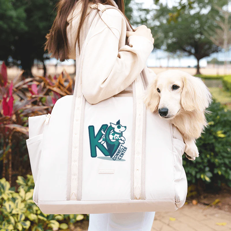 A woman walks with a dog in a tote bag with a K9 Connect Center Logo on it.