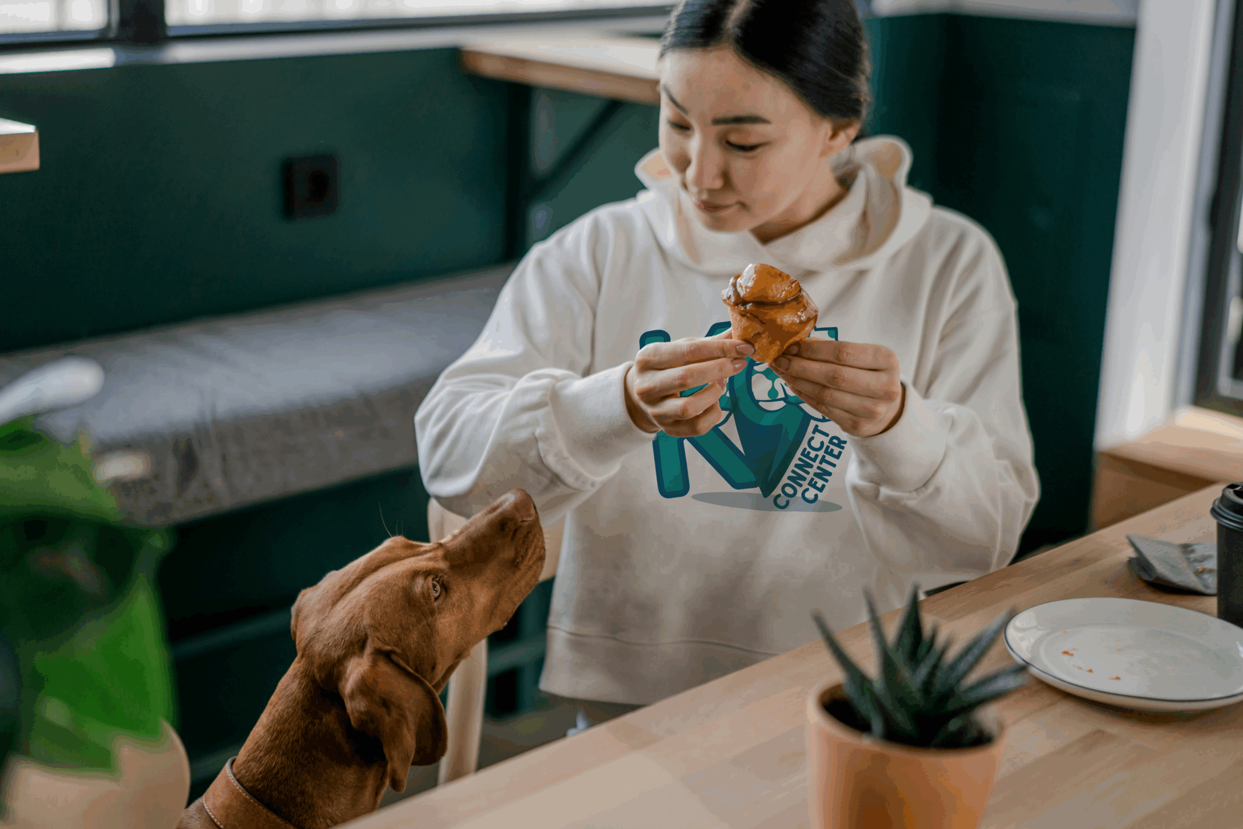 A woman wearing a K9 Connect Center logo hoodie eats a donut while her dog begs for a bite.