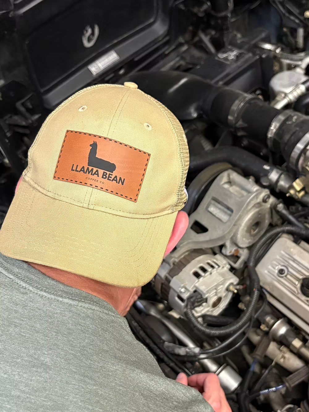 Man wears a Llama Bean Coffee Co hat while bending over a car engine